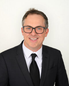 Man with dark rim glasses in black suit and tie and white shirt