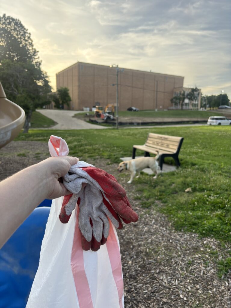 Trash bag, work gloves being held in a hand, with a dog in the background