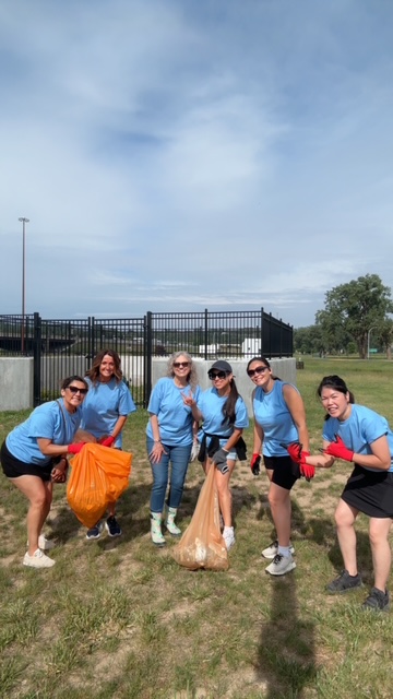 Group of 7 realtors in blue shirts posing with smiles while picking up trash in orange bags