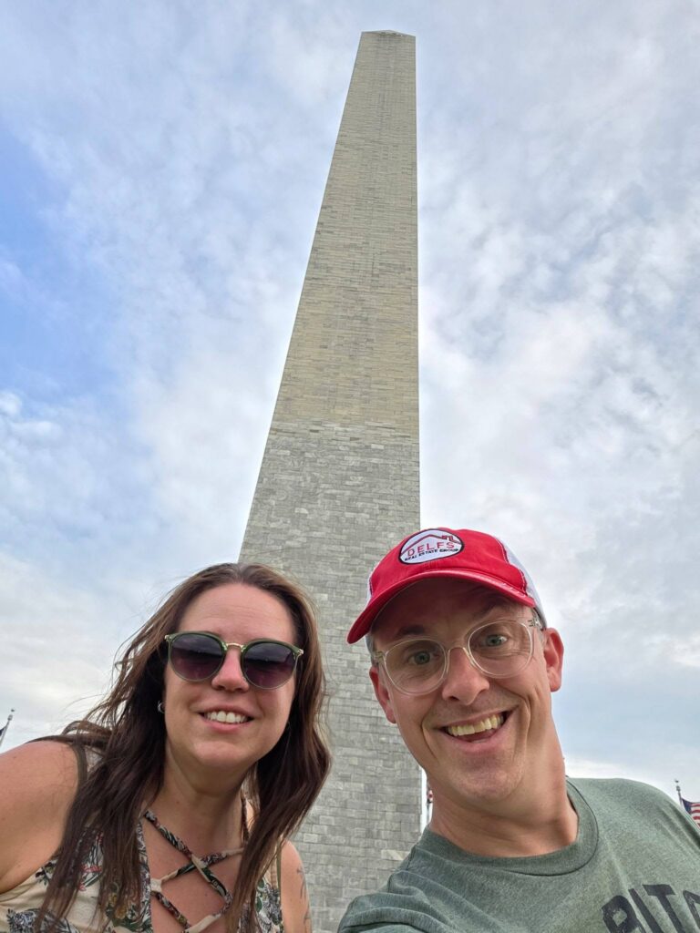 Denise Gutierrez in sunglasses and Chris Delfs in red hat stand outside the Washington Monument looking happy and excited on a sunny day with clear blue skies
